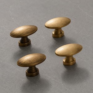 May include: Four oval-shaped cabinet knobs with a brushed antique brass finish. Each knob features a smooth, rounded top surface and a tapered cylindrical base, showcasing a consistent metallic texture and warm golden-brown color throughout.