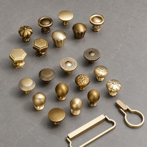 May include: A collection of various brass-toned cabinet knobs and pulls in diverse shapes and textures, including smooth spheres, fluted designs, hexagonal forms, and intricate patterns, arranged together in a group on a flat surface.