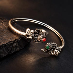 May include: A silver-toned open cuff bracelet featuring two detailed elephant motifs at the ends. One elephant is accented with a small, round red stone, while the other features a green stone. The metal has a polished finish with textured details.