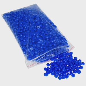 May include: A clear plastic resealable bag filled with a large quantity of small, round, solid blue wax beads. Some of the blue wax beads have spilled out from the open top of the bag, creating a small pile on the surface below the container.