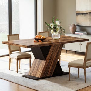 May include: A rectangular dining table with a dark wood grain finish and a geometric V-shaped pedestal base. The table is accompanied by two chairs featuring light beige upholstered seats and backs, with a bowl of oranges and a vase of white flowers.