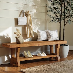 May include: A rectangular wooden bench with a lower shelf, featuring a natural brown finish. Two white patterned throw pillows sit on the bench, while a pair of white heels and white slippers are placed on the lower shelf for storage.