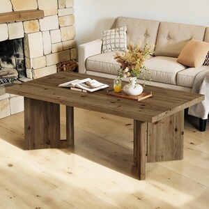 May include: A rectangular wooden coffee table with a natural brown finish and visible wood grain, featuring a thick tabletop and two sturdy vertical slab legs. An open book, a small glass of yellow liquid, and a white vase with dried plants sit on top.