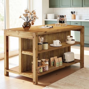 May include: A rectangular wooden kitchen island with a natural grain finish, featuring an open shelving unit on one side. The top surface holds a glass vase with white flowers and a magazine. Shelves contain white mugs, bowls, jars, and a toaster.