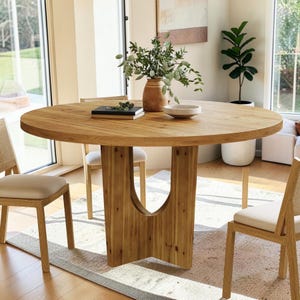 May include: A round light brown wooden dining table with a central pedestal base featuring a cutout design. The table is surrounded by wooden chairs with light-colored upholstered seats. A ceramic vase with green foliage sits on the tabletop.