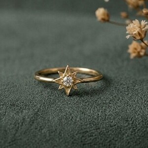 Op de afbeelding: Een gouden ring met een centrale stervormige zetting met een kleine, heldere, rondgeslepen edelsteen. De ringband is dun en gepolijst en heeft een heldere metalen afwerking die contrasteert met het ingewikkelde, puntige stervormige ontwerp.