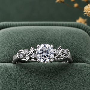 Puede incluir: Un anillo de tono plateado con una gran piedra central redonda de talla brillante flanqueada por piedras más pequeñas. La banda presenta un intrincado diseño de metal con formas de hojas y enredaderas en ambos lados.