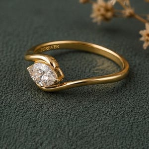 May include: A gold ring featuring a marquise-cut clear gemstone set in a bypass-style band. The inner surface of the gold band is engraved with the word FOREVER in a clear, sans-serif font, visible along the upper curve of the ring's shank.