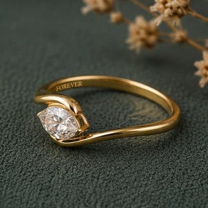 May include: A gold ring featuring a marquise-cut clear gemstone set in a bypass-style band. The inner surface of the gold band is engraved with the word FOREVER. The ring has a polished finish and a slender, curved design that holds the stone.