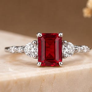 Peut inclure: Une bague de couleur argentée avec une grande pierre précieuse rouge de taille émeraude rectangulaire au centre. La bague est flanquée de deux pierres rondes, claires et taillées en brillant de chaque côté, avec des pierres plus petites sur l'anneau.