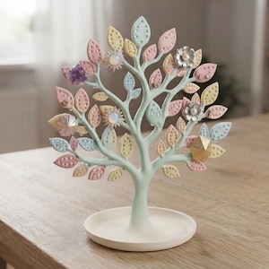 May include: A light mint green jewellery stand shaped like a tree with a circular base. The branches feature numerous leaves in pastel shades of pink, yellow, and blue, each with small perforations. Several rings are displayed on the branches.