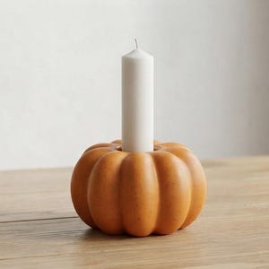 May include: A matte orange pumpkin-shaped candle holder with a smooth, rounded, ribbed surface. A single, tall, cylindrical white taper candle is inserted into the centre opening of the pumpkin base, standing upright as a singular decorative unit.