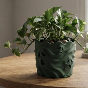 May include: A dark green cylindrical planter with a matte finish and a wavy, draped texture featuring embossed ghost faces. A trailing pothos plant with variegated green and white heart-shaped leaves grows from the top of the container.