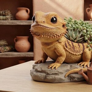 May include: A yellow and brown bearded dragon figurine planter featuring large black eyes and textured scales. The lizard is perched on a gray rock base with a succulent plant growing from its back and a small brown watering can placed nearby.