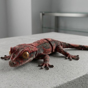 Könnte beinhalten: Eine realistische, bewegliche Gecko-Figur mit strukturiertem Körper in einem gesprenkelten Rot- und Dunkelgrau-Muster. Die Eidechse hat große, markante goldbraune Augen mit vertikalen Pupillen und sichtbare Gelenke an Rücken und Gliedmaßen.