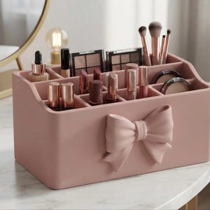 May include: A dusty pink rectangular makeup organiser featuring a prominent moulded bow on the front. The container has multiple compartments of varying sizes, currently holding assorted lipsticks, makeup brushes, compacts, and a small glass bottle.
