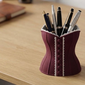 May include: A burgundy pen holder shaped like a corset, featuring a vertical row of matching fabric buttons down the center, white scalloped trim along the edges, and a collection of various metal pens stored inside the open top opening.