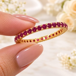 May include: A thin gold eternity band featuring a continuous row of small, round, vibrant pinkish-red gemstones set in a shared-prong setting. The metal band has a polished finish, and the stones are uniform in size and color throughout the ring.