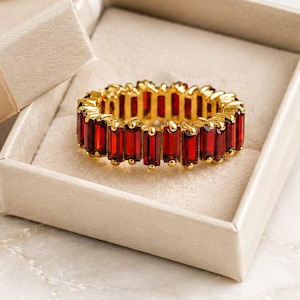 May include: A gold eternity ring featuring a continuous row of rectangular, deep red baguette-cut gemstones set in a metal band, presented inside an open, light beige square gift box with a matching satin ribbon tied in a bow on the lid.