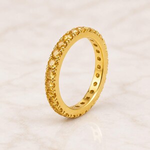 Puede incluir: Un anillo de tipo alianza en oro con una hilera continua de piedras preciosas de citrino amarillo de talla redonda, engastadas en un marco de metal dorado pulido. El anillo presenta un diseño uniforme con piedras amarillas brillantes.