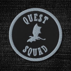 May include: A circular embroidered patch featuring a black background with a white border. The text QUEST SQUAD is stitched in white capital letters, arching above and below a central white silhouette of a flying dragon with spread wings.