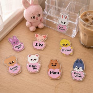 Puede incluir: Una colección de ocho dijes de acrílico transparente con personajes de animales de dibujos animados que sostienen corazones rosas con nombres impresos en negro: Chang Bin, I.N, Felix, Seung min, Hyun Jin, Han, Bang Chan y Lee Know.