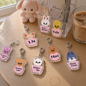 Puede incluir: Una colección de ocho llaveros de acrílico transparente con personajes animales de dibujos animados que sostienen corazones rosas. Cada corazón tiene un nombre impreso: Lee Know, Felix, I.N, Chang Bin, Seung min, Hyun Jin, Han y Bang Chan.