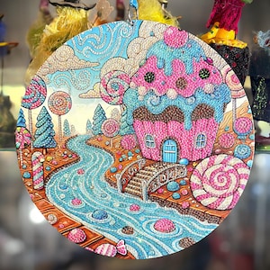 May include: A round, colorful ornament featuring a whimsical candy-themed landscape. The artwork depicts a pink cupcake house, a flowing blue river, and lollipop trees. The ornament is embellished with intricate details and vibrant colors.