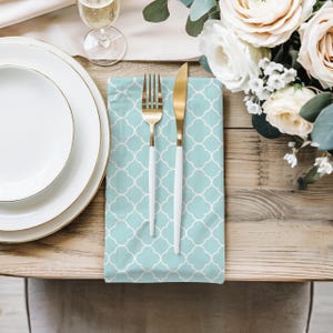 May include: A light blue cloth napkin featuring a white quatrefoil pattern, topped with a gold-toned metal fork and a knife with a gold-toned metal blade and a white handle, arranged neatly on a wooden surface next to white plates and flowers.