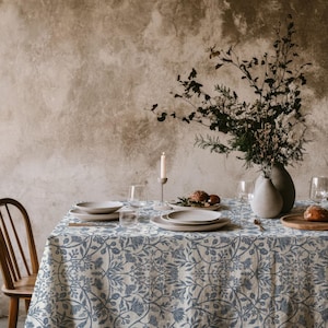 May include: A dining table covered with a light-coloured tablecloth featuring a blue floral pattern. The table is set with white ceramic plates, clear glassware, a single white taper candle in a holder, and a vase filled with dried branches.