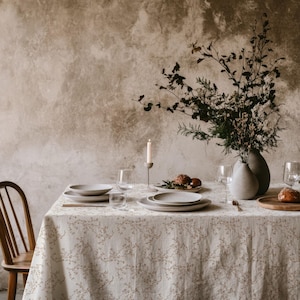 Op de afbeelding: Een eettafel bedekt met een beige linnen tafelkleed met een delicaat bruin bloemenpatroon. De tafel is gedekt met keramische borden, helder glaswerk, een enkele dinerkaars in een houder en een vaas gevuld met gedroogde takken.