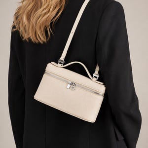 May include: A structured, rectangular cream-colored handbag with a textured finish. The bag features a top handle, a long adjustable shoulder strap, and a silver-toned metal zipper closure that runs along the top perimeter of the main compartment.