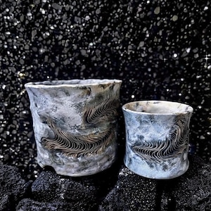 Może przedstawiać: Dwa cylindryczne naczynia ceramiczne o teksturowanym, szaro-białym wykończeniu. Każdy element posiada wyraźny, wypukły poziomy pas ze skomplikowanymi, ciemnobrązowymi, falistymi wzorami liniowymi na zewnętrznej powierzchni.