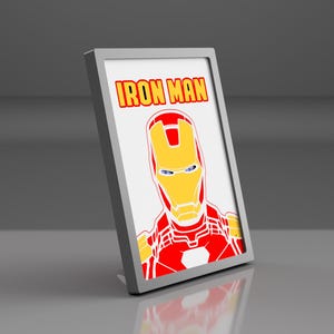 Peut inclure: Un cadre rectangulaire de couleur argentée contenant une impression graphique du personnage Iron Man. Le personnage est représenté dans une armure rouge et jaune sur fond blanc, avec le texte IRON MAN imprimé en lettres jaunes en haut.