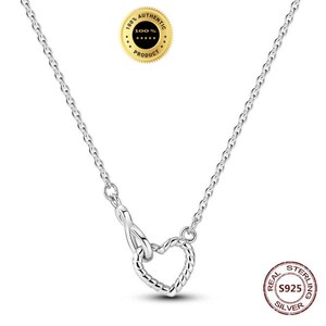 May include: A silver-coloured chain necklace featuring a heart-shaped pendant with a twisted rope texture. The necklace includes a lobster claw clasp mechanism. A gold-coloured circular seal with the text 100% AUTHENTIC PRODUCT and a stamp reading REAL STERLING S925 SILVER are visible.