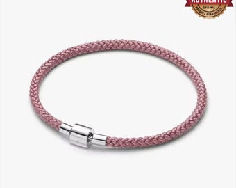 May include: A mauve braided cord bracelet featuring a polished silver-tone cylindrical clasp. The bracelet has a circular shape and a textured woven surface. A circular gold-toned badge in the upper right corner displays the text 100% AUTHENTIC SILVER.