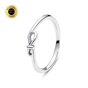 Könnte beinhalten: Ein polierter silberfarbener Ring mit einem zarten, unendlichkeitsförmigen Knotenmuster auf einer Seite. Das Band ist glatt und kreisförmig und weist eine reflektierende metallische Oberfläche auf. Ein goldenes Siegel mit dem Text 100% AUTHENTIC PRODUCT ist sichtbar.