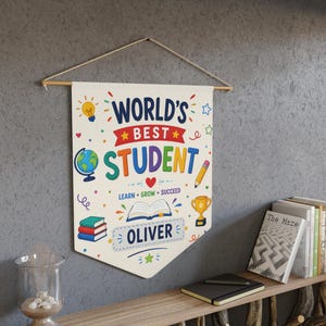 Könnte beinhalten: Ein cremefarbenes Wandbanner aus Stoff mit dem Text "WORLD'S BEST STUDENT" in bunten Buchstaben sowie Illustrationen einer Glühbirne, eines Globus, eines Bleistifts, eines offenen Buches, eines Pokals und eines Bücherstapels. Der Name "OLIVER" steht in einem blauen Kasten.