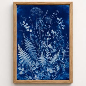 May include: A framed cyanotype print featuring a collection of white botanical silhouettes, including ferns and wildflowers, set against a deep, saturated navy blue background. The artwork is encased in a light-toned wooden frame with a clean finish.