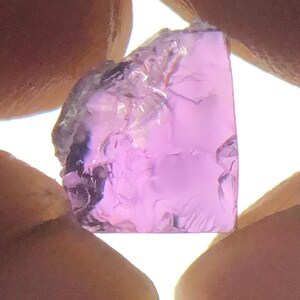 May include: A translucent, light purple gemstone with an irregular, faceted shape. The stone displays a vibrant lavender hue and exhibits a natural, raw texture with visible internal inclusions and sharp, jagged edges throughout its surface.