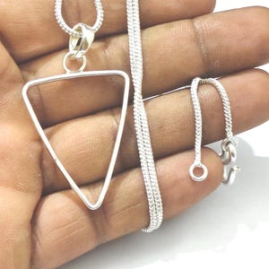 Op de afbeelding: Een zilverkleurige ketting met een holle driehoekige hanger aan een fijne, getextureerde ketting. De ketting is voorzien van een klein rond sluitingsmechanisme aan het uiteinde en heeft een metallic afwerking met een geometrisch ontwerp.
