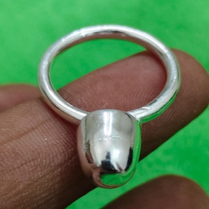 May include: A polished silver ring featuring a smooth, rounded, teardrop-shaped accent piece attached to a circular band. The metal surface is reflective and shiny, showcasing a clean, minimalist design with a solid, continuous metallic construction.