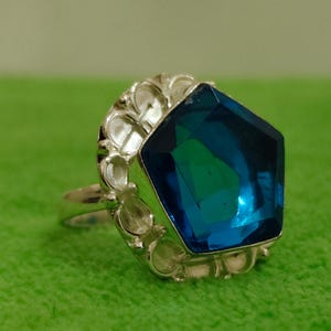 May include: A silver-toned ring featuring a large, pentagonal, faceted blue gemstone set within a decorative, scalloped metal frame. The ring band is thin and metallic, supporting the prominent, deep blue stone that reflects light across its surface.