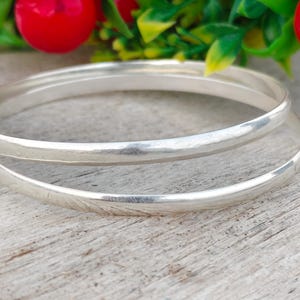 Op de afbeelding: Twee gladde, gepolijste zilveren armbanden met een afgerond profiel. De armbanden zijn cirkelvormig en hebben een reflecterende, metalen afwerking. Deze eenvoudige, onversierde sieraden worden als paar gepresenteerd met een strak ontwerp.