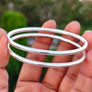 May include: Two smooth, polished silver-toned bangle bracelets with a rounded profile. The bangles feature a continuous, seamless circular design with a reflective metallic finish, showcasing a simple and clean aesthetic for wrist adornment.