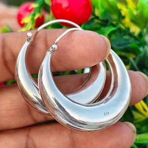 May include: A pair of polished silver hoop earrings with a thick, rounded profile. The earrings feature a smooth, reflective surface and a hinged post closure. A small hallmark stamp reading 925 is visible on the lower curve of the metal hoop.