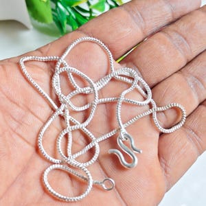 May include: A silver-colored chain necklace featuring a textured, rope-like link design. The necklace includes a distinct S-shaped hook clasp at one end and a small circular jump ring at the other end, allowing for a secure and adjustable closure.