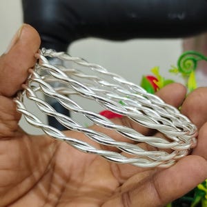 May include: A set of four silver-colored bangles, each featuring a twisted, rope-like design. The bracelets have a polished, reflective metallic finish and are circular in shape, showcasing a consistent spiral pattern throughout the entire band.