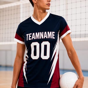 Puede incluir: Una camiseta de cuello en V azul marino con ribetes blancos en el cuello y las mangas. El frente presenta el texto TEAMNAME en letras mayúsculas blancas sobre el número 00. Los costados tienen acentos gráficos angulares granates y blancos.