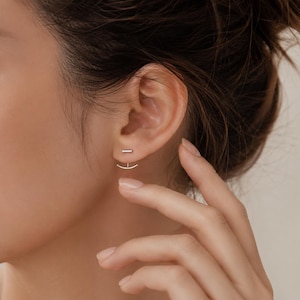 Peut inclure: Une boucle d'oreille de type ear jacket argentée composée d'un petit clou en forme de barre horizontale sur le lobe et d'une pièce arquée incurvée qui pend sous le lobe, créant un effet superposé avec une finition métallique polie.